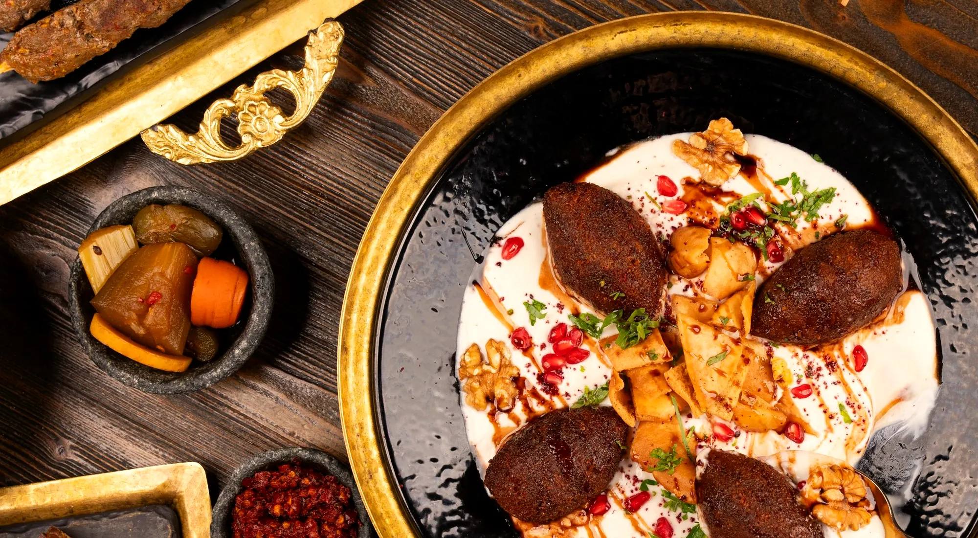 Kibbeh in warm yoghurt, lamb kofta skewers, pickles and toasted bread on a dark wooden table