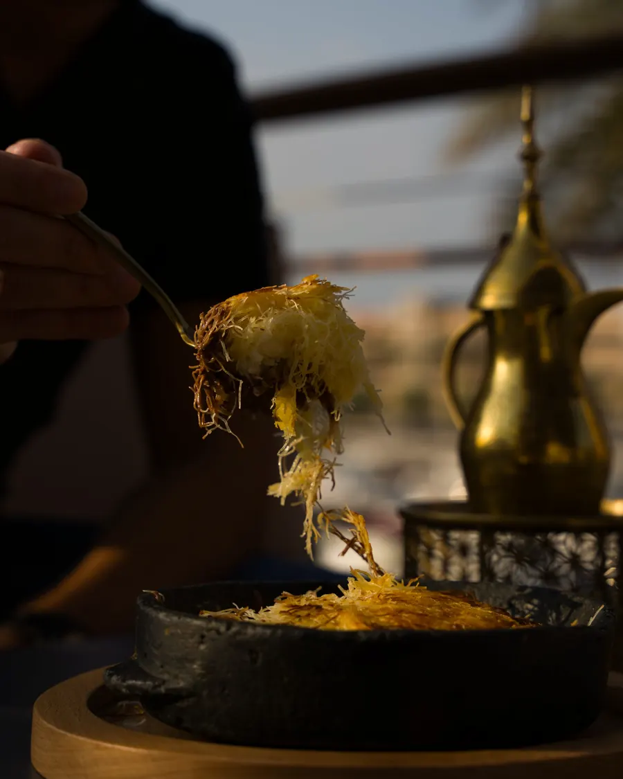 A forkful of warm knafeh lifted from a cast-iron pan, a brass coffee pot behind
