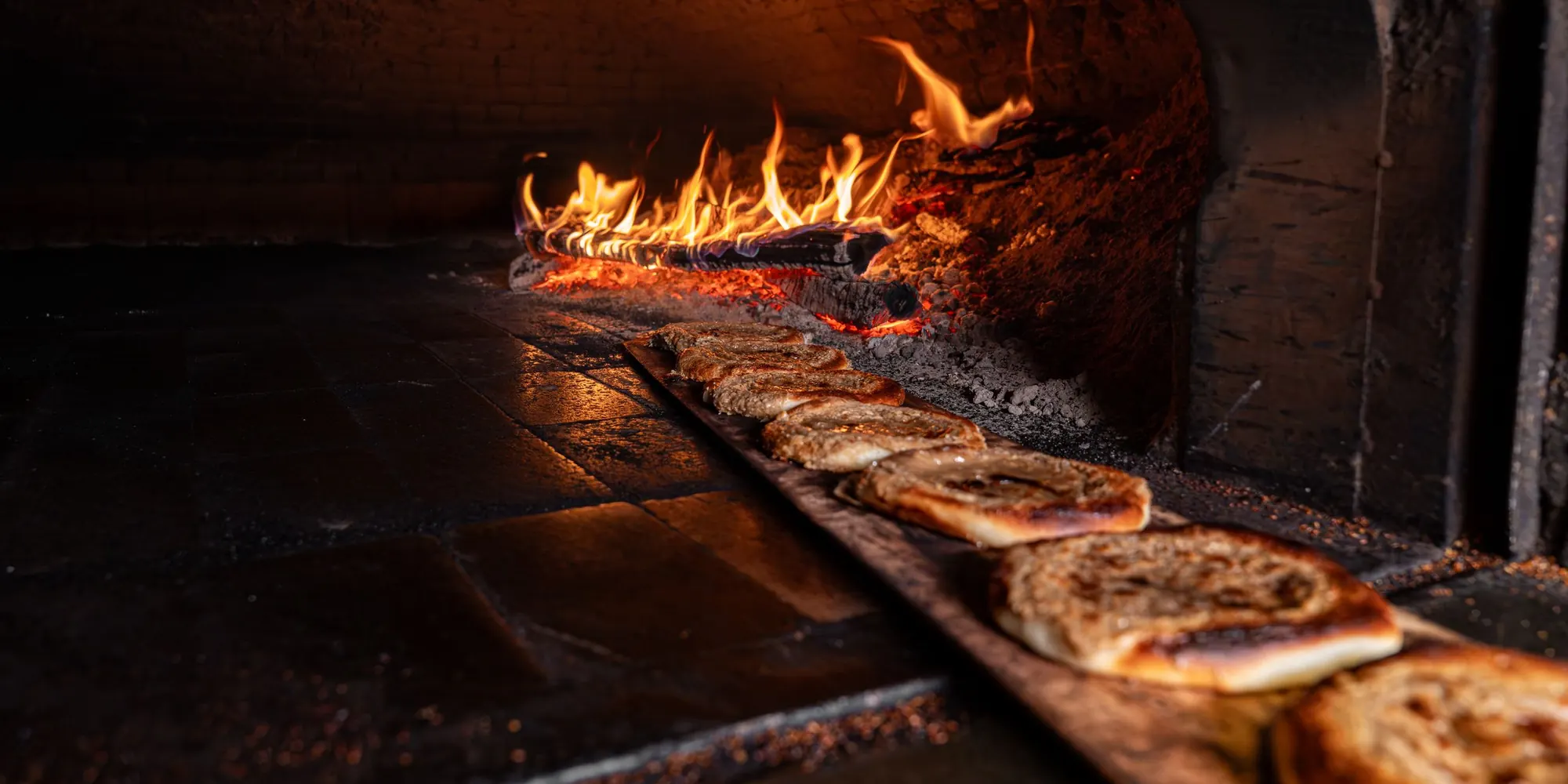 Flatbreads lined up on a wooden peel beside the flames of a wood-fired oven